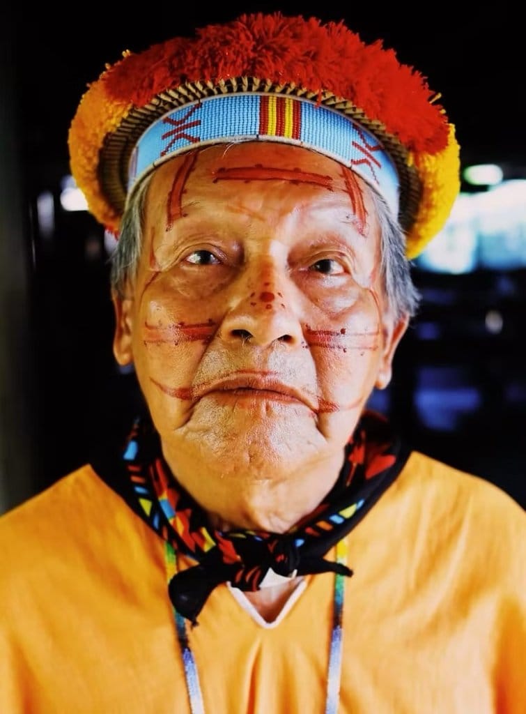Basilio, an indigenous Amazonian elder, wearing traditional red face paint and a beaded headband with red pom-pom headdress