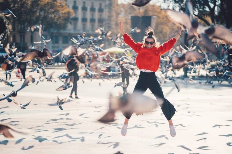 Woman in a red sweater jumping with arms raised in a city plaza as a flock of pigeons takes flight around her