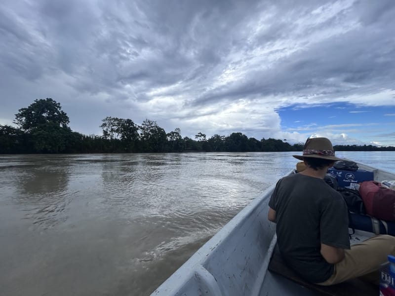 Ben Hoffman riding in a canoe on a muddy Amazon river in Ecuador, dense jungle lining both banks under a dramatic overcast sky