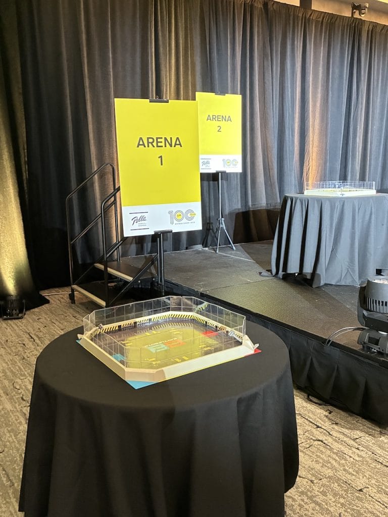 Battle Bots event setup with yellow Arena 1 and Arena 2 signs on a stage and a miniature octagonal battle arena on a table in the foreground