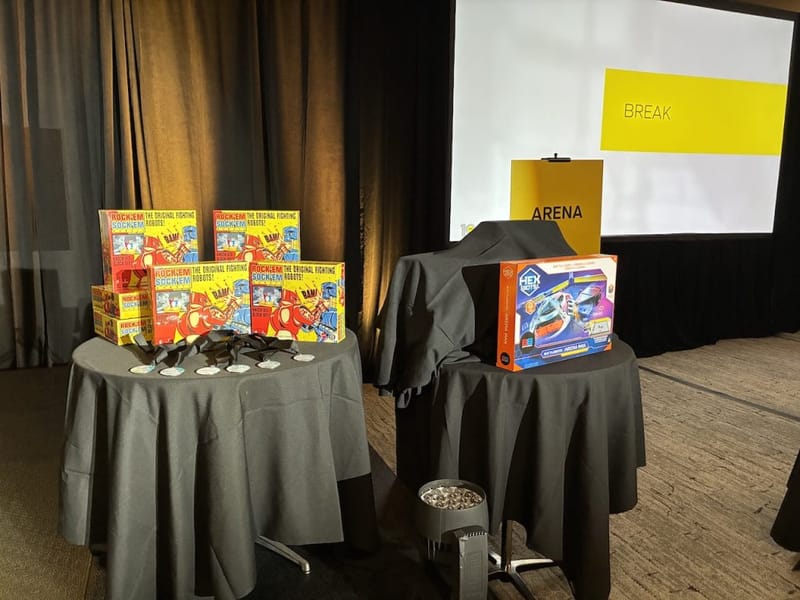 Rock Em Sock Em Robots game boxes and a Hex Bugs Battlebots Arena Max kit displayed on tables at a Battle Bots team-building event venue