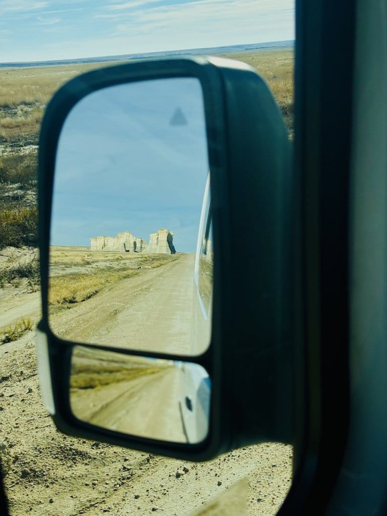 Vehicle side mirror reflecting a dirt road leading toward chalk rock formations on the open plains