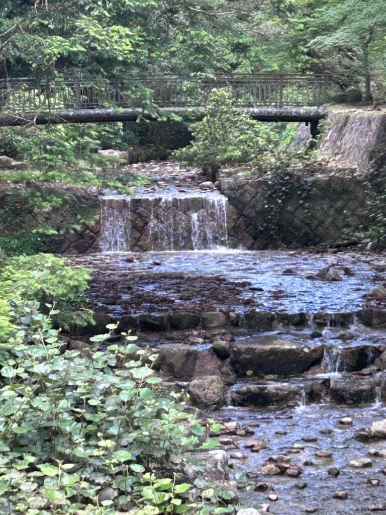 Small waterfall cascading over rock steps in a forested Japanese garden, with a metal footbridge in the background