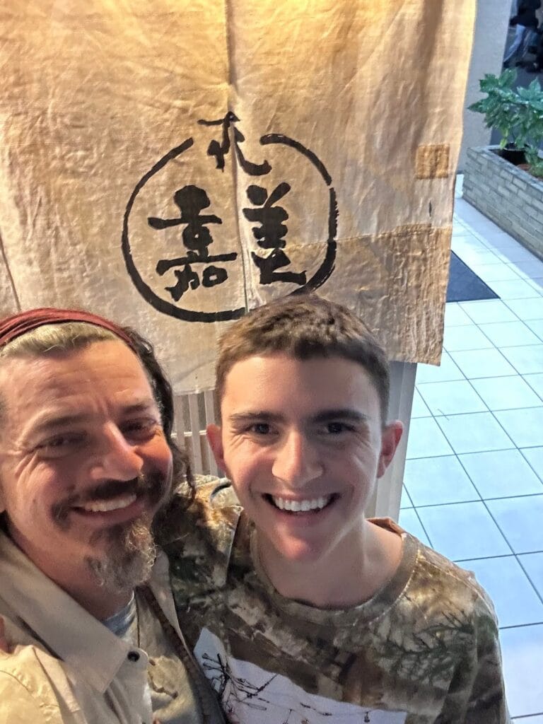 Ben Hoffman and a travel companion smiling in front of a Japanese noren curtain at a restaurant entrance in Japan