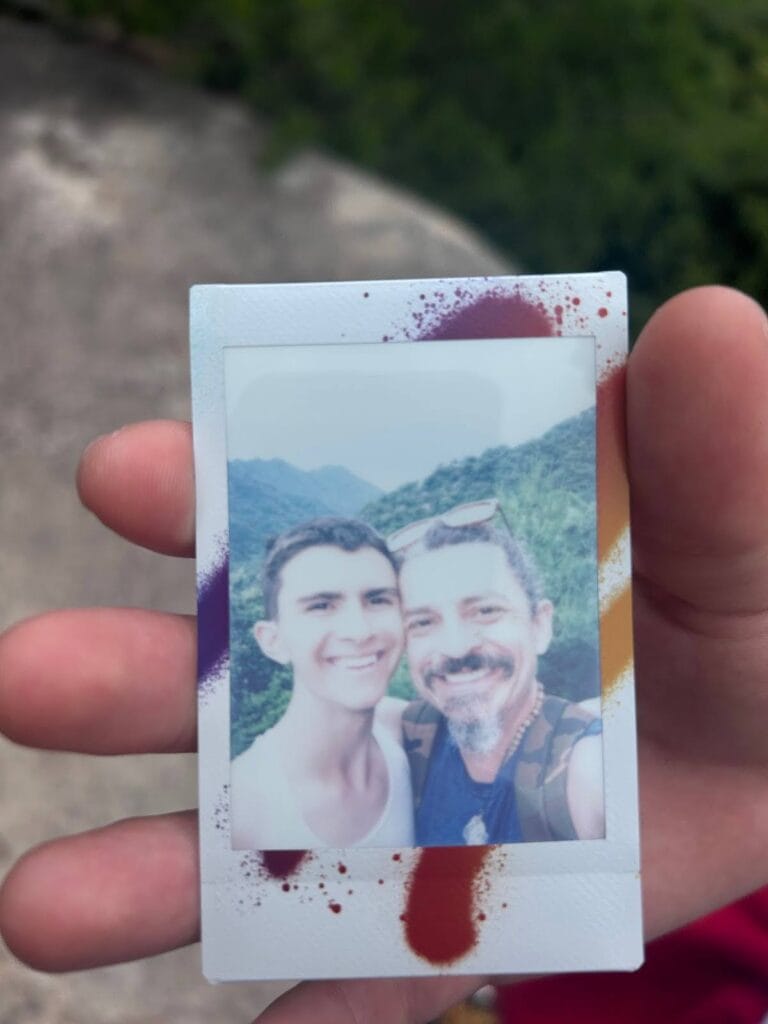 Hand holding a paint-splattered Instax photo of Ben Hoffman and a companion smiling in front of green mountains in Japan