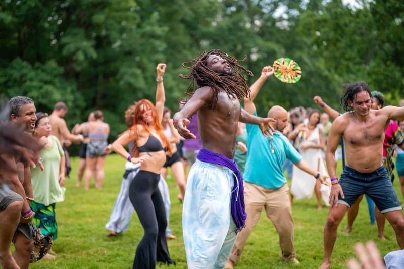 Crowd of people dancing outdoors at a Sol Dance event, with a shirtless man in white pants and purple sash in the foreground moving with arms outstretched and eyes closed