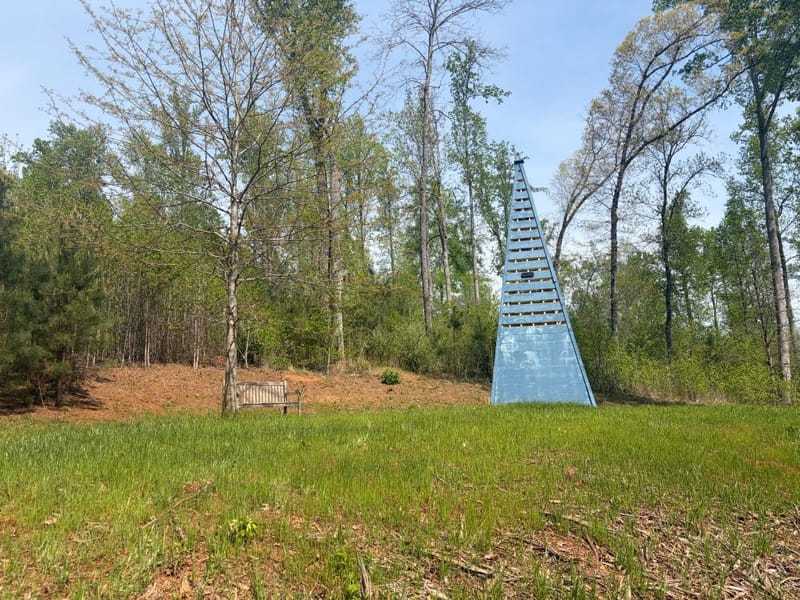 Tall blue pyramid structure in a wooded clearing at Serenbe, with a small bench along the tree line