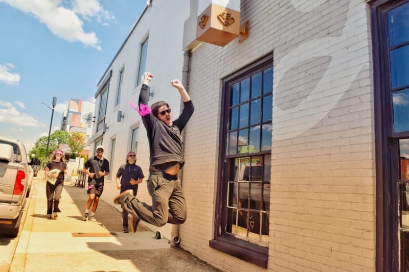 Man jumping with fists raised in celebration on a Nashville sidewalk, with three teammates running behind him past a white brick building