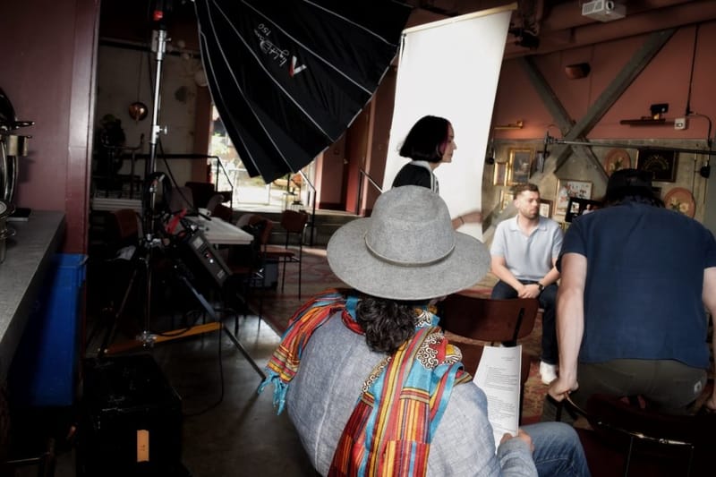 Behind-the-scenes of a video production shoot in Nashville, with a softbox light rig, a crew member in a wide-brimmed hat reviewing a script in the foreground, and a man seated as the interview subject