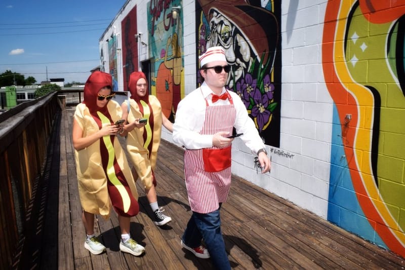 Two participants in hot dog costumes and a game leader in a striped apron walking past colorful Nashville street murals during a cityHUNT scavenger hunt