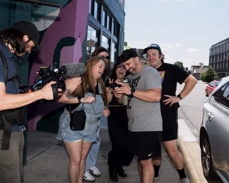 Group of excited people gathered around someone checking a phone on a Nashville sidewalk, videographer filming nearby, colorful street mural in the background