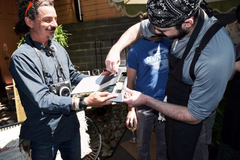Man with an instant camera around his neck sharing a photo album with a bearded guest at an outdoor Nashville gathering